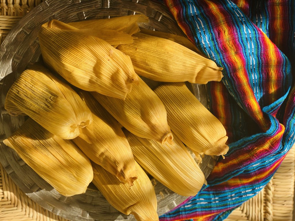 Tamales with corn husks on a pewter platter with a colorful Mexican reboso.