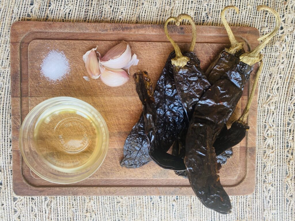 Wooden cutting board with pasilla chiles, garlic cloves, salt and bowl of oil