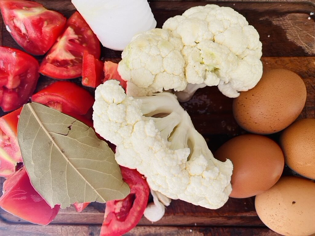 Mexican cauliflower fritter ingredients on a cutting board, cauliflower chunks, eggs, onion, tomatoes, and bay leaf