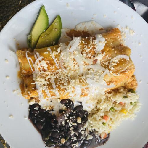 Enchiladas Suizas Rojas on a white plate with beans, rice, and avocado slices
