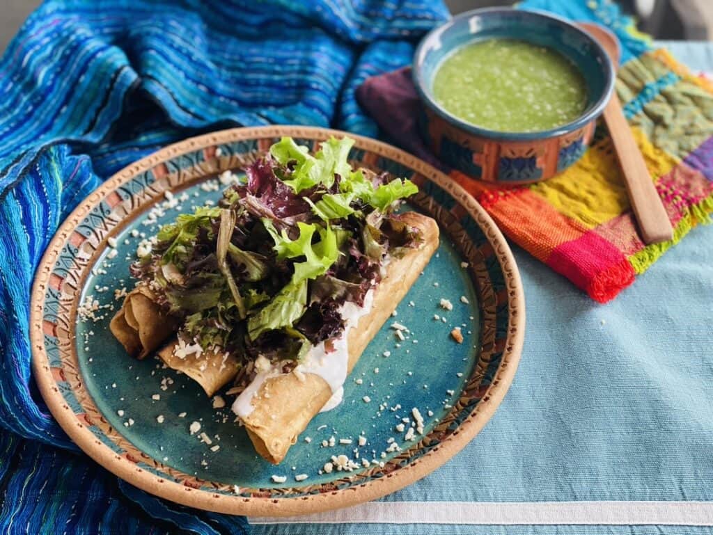 3 Tacos dorados on a blue potter plate, topped with lettuce, sour cream, shredded cheese and accompanied by a bowl of salsa verde
