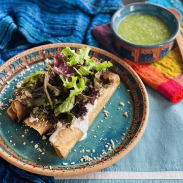 3 Tacos dorados on a blue potter plate, topped with lettuce, sour cream, shredded cheese and accompanied by a bowl of salsa verde