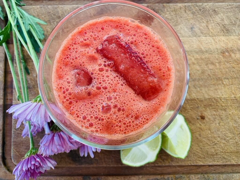 Watermelon ginger limeade in a glass with ice cubes, sitting on top of a wooden cutting board, purple flowers and lime wedges are on the side