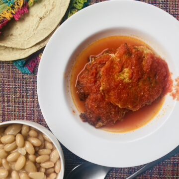 A white bowl with 2 chicken and vegetable fritters in tomato sauce, with a small bowl of Peruvian beans on the side and 2 tortillas in a Mexican-style napkin