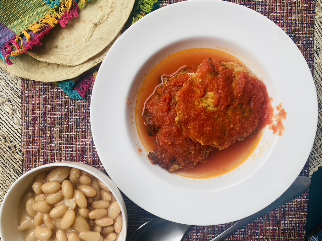 A white bowl with 2 chicken and vegetable fritters in tomato sauce, with a small bowl of Peruvian beans on the side and 2 tortillas in a Mexican-style napkin