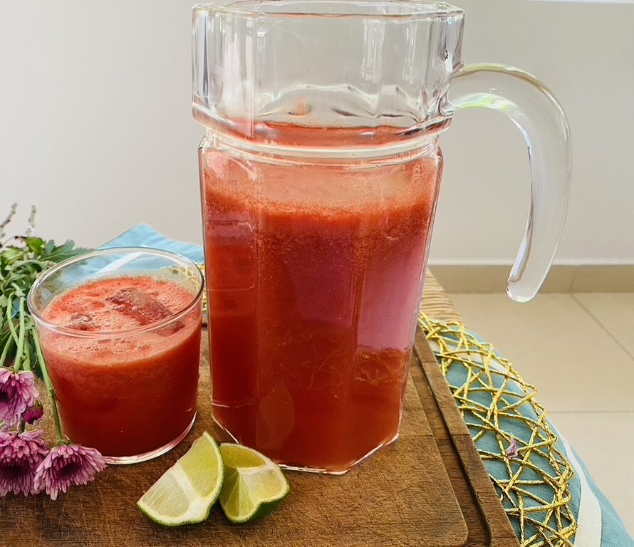 Pitcher and glass of watermelon ginger limeade sitting on top of a wooden cutting board, with lime wedges
