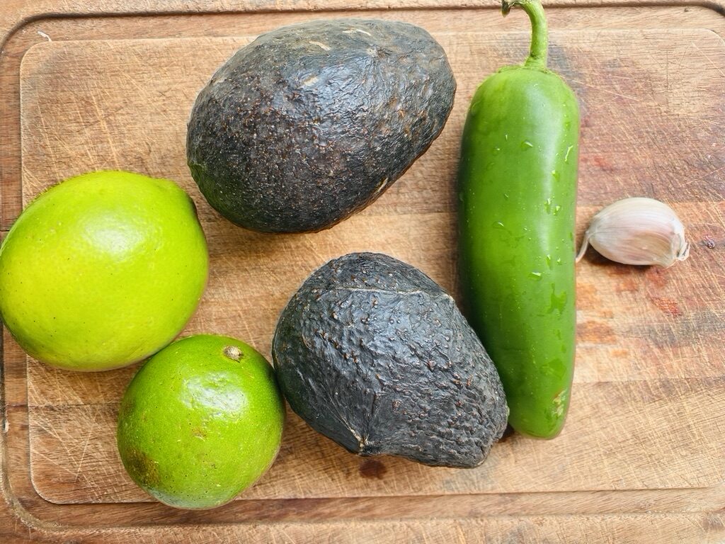 Cutting board with creamy avocado salsa ingredients: limes, avocados, jalapeño pepper and garlic clove