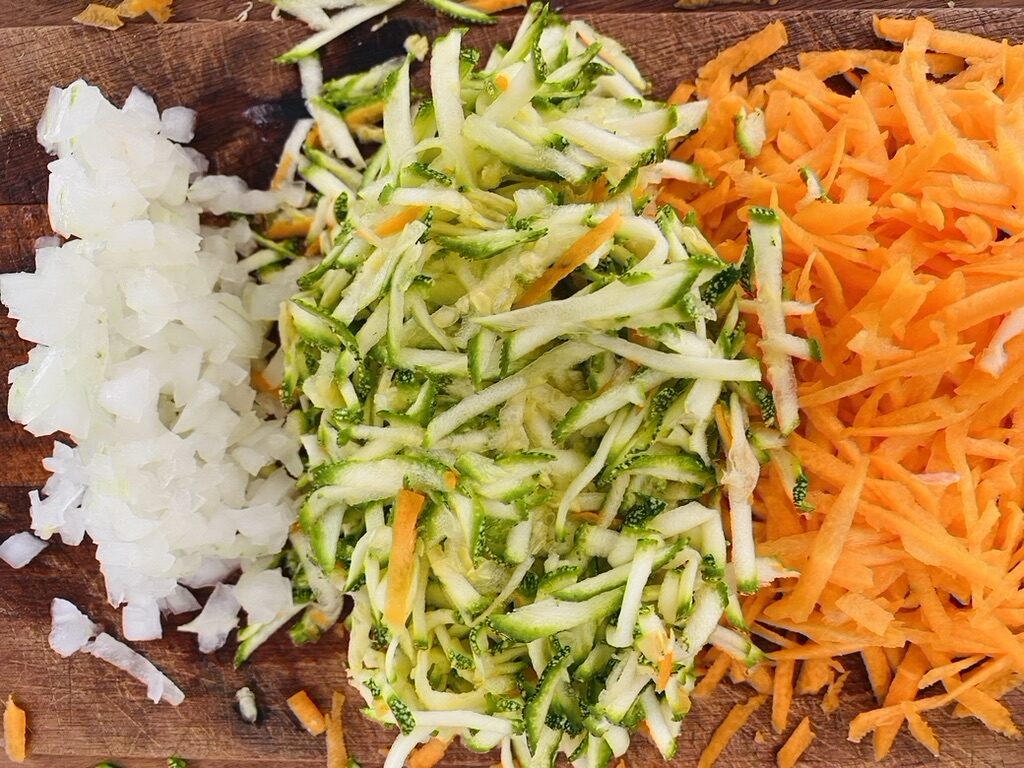 A wooden cutting board with diced white onion, shredded Mexican grey squash, and shredded carrots