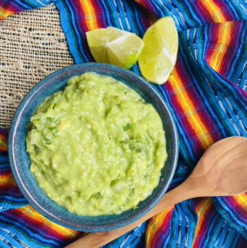 Creamy avocado and tomatillo salsa in a blue pottery bowl with a wooden spoon and lime wedges on the side.