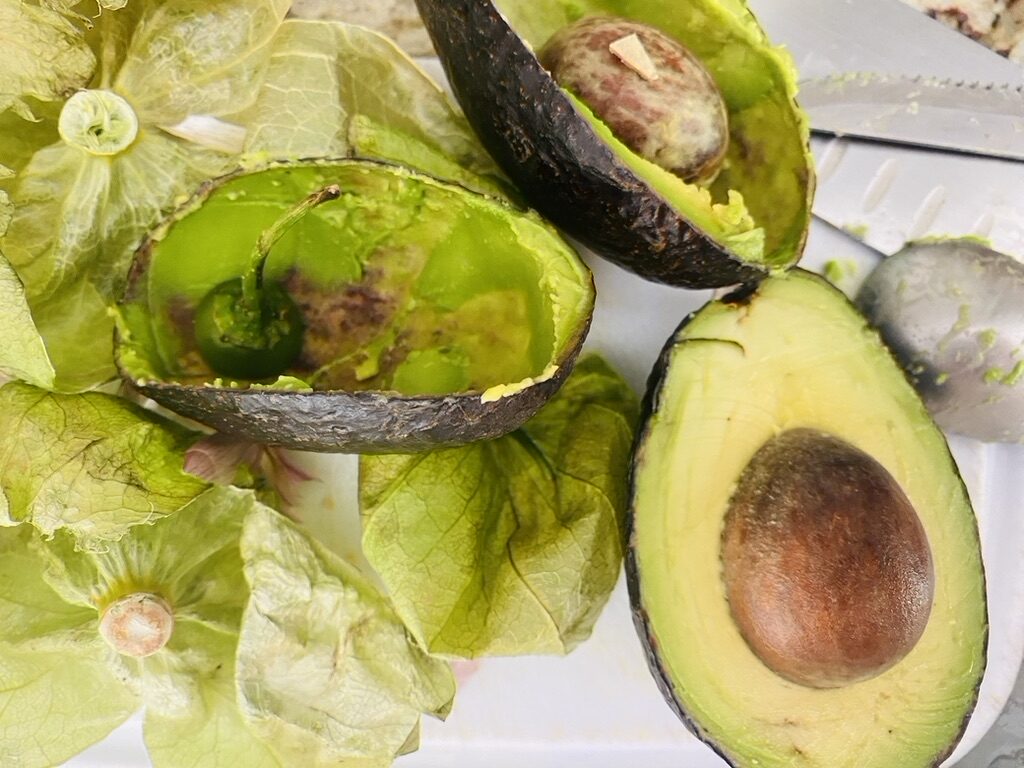 White cutting board topped with tomatillo husks, avocado peels, and a half of an avocado with the pit