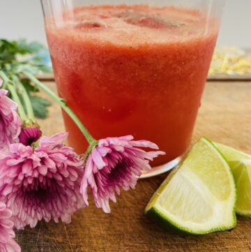 Glass of watermelon ginger limeade sitting on a wooden board with lime wedges and purple flowers on the side