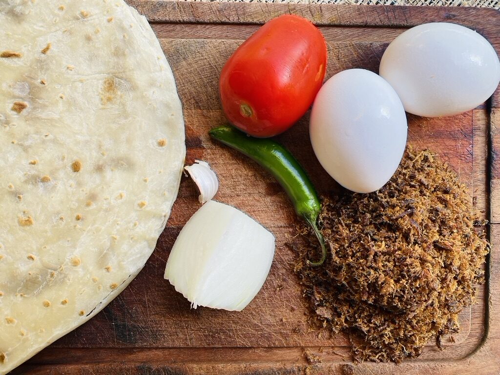 Wooden cutting board with machaca, tomato, onion, chile pepper, flour tortillas, eggs, and garlic clove