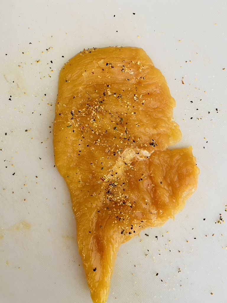 Chicken milanesa being seasoned with salt, pepper, and garlic powder on a white cutting board