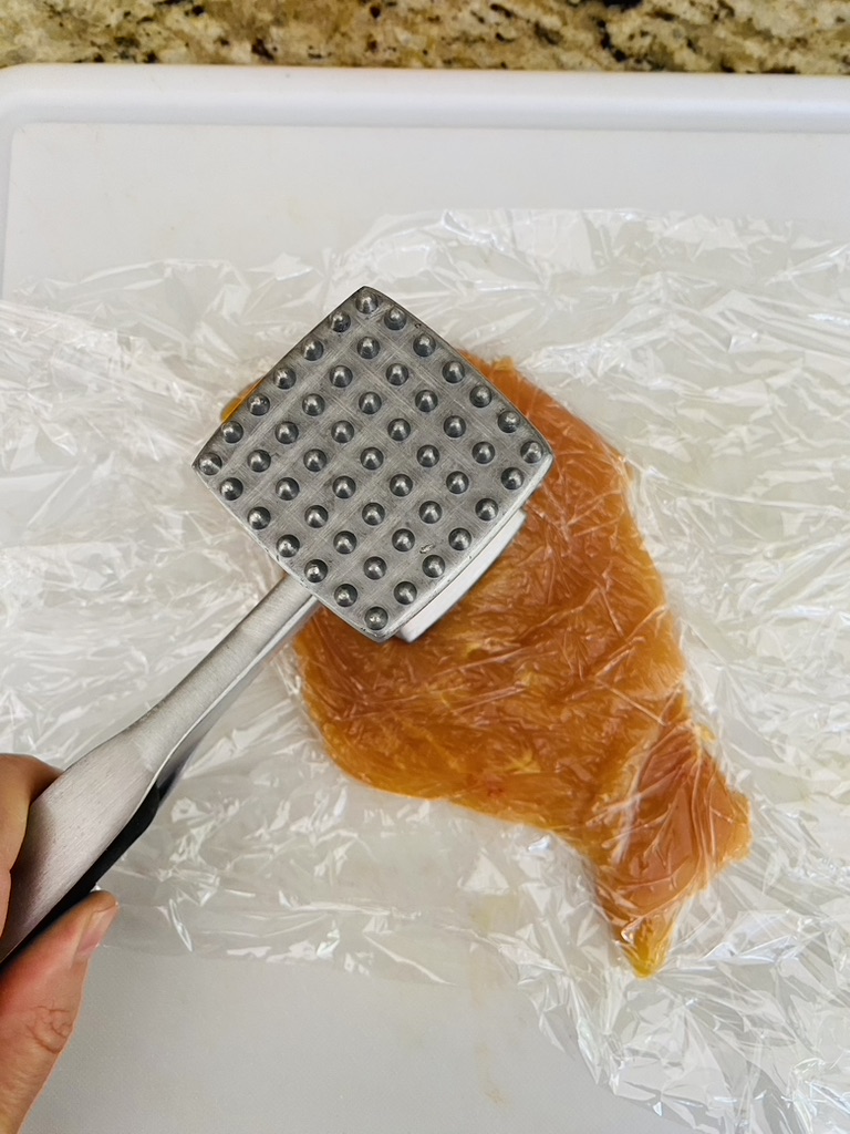 Chicken cutlet between 2 sheets of plastic wrap on a cutting board being pounded thin with a meat mallet