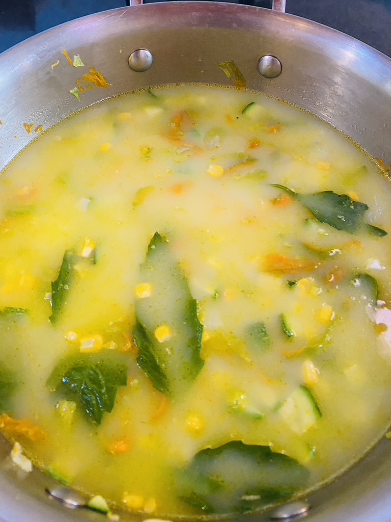 Metal soup pan with squash blossoms, onion, diced squash, corn, broth, and epazote leaves