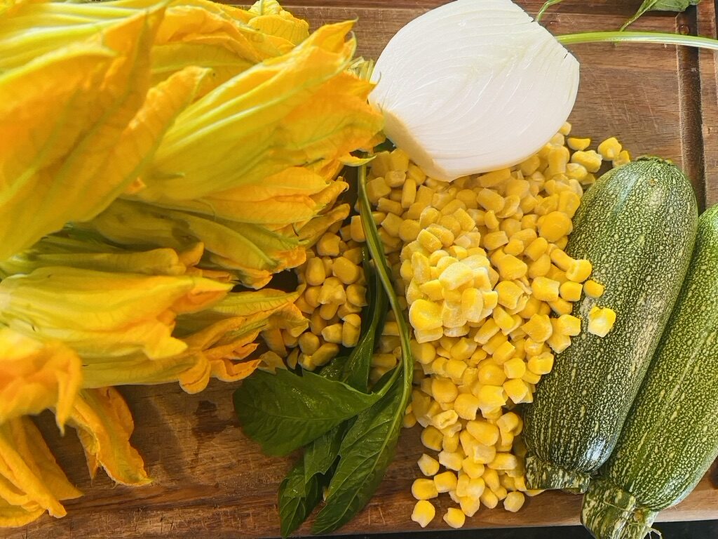 Wooden cutting board with squash blossoms, corn kernels, epazote stem, 2 Mexican grey squashes, and 1/2 onion