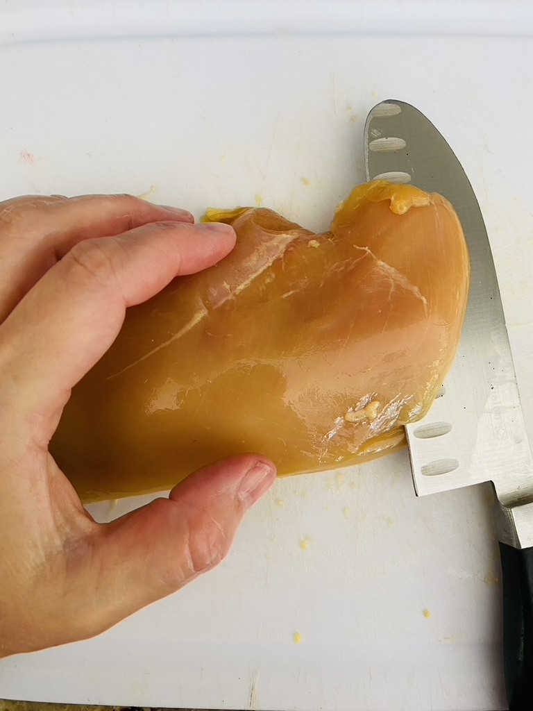Whole boneless, skinless chicken breast on a white cutting board being sliced through the center