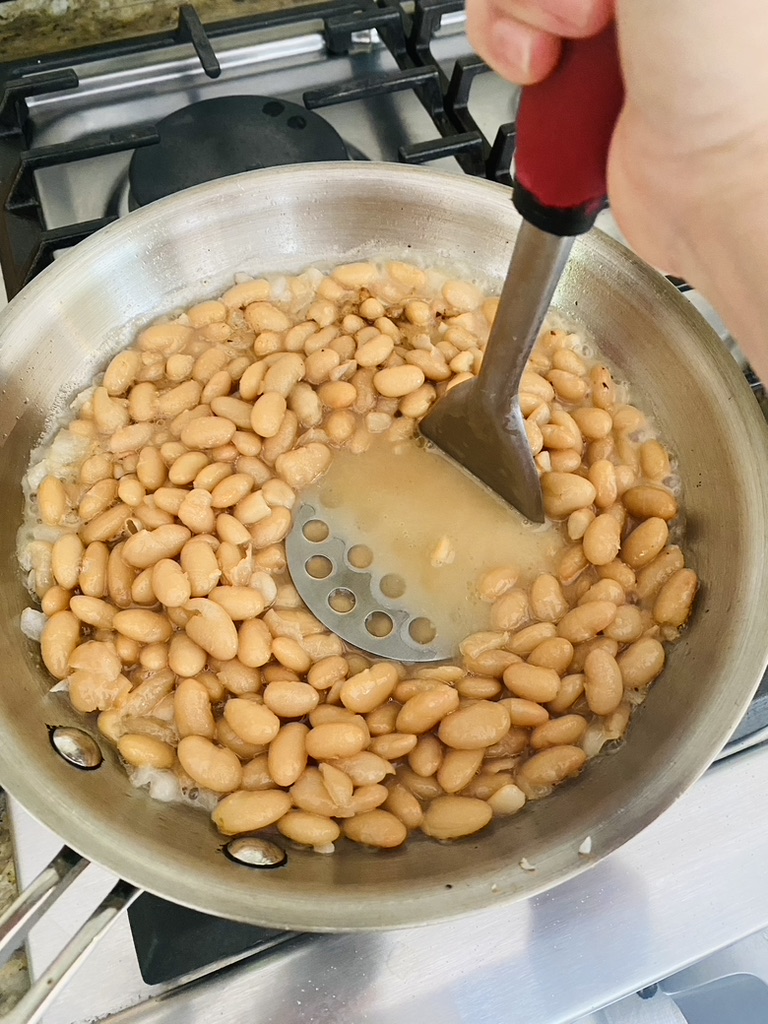 Beans in a skillet being mashed with a potato masher