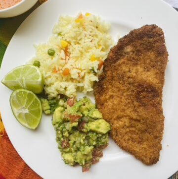 Chicken milanesa on a white plate with a side of guacamole, white rice, and lime wedges