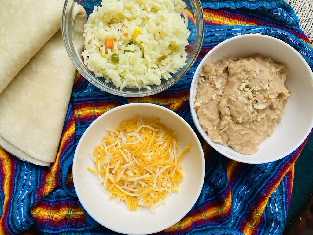 Flour tortillas, bowl of Mexican white rice, bowl of refried beans, and cheese on a zolorful Mexican cloth