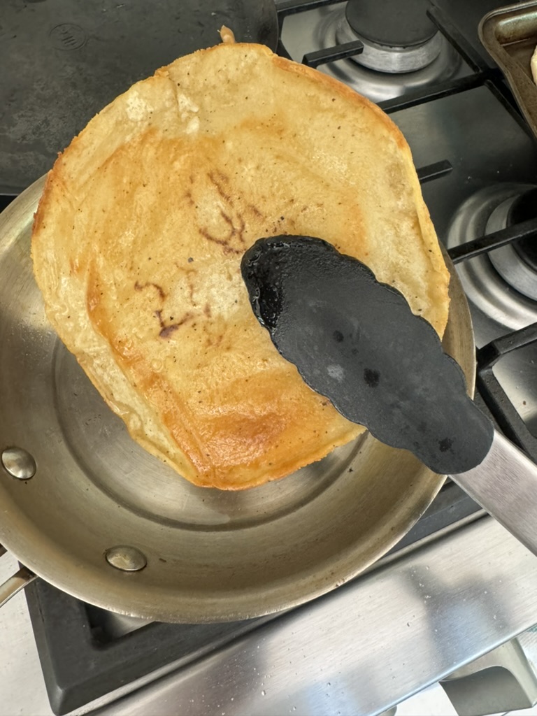 Golden fried corn tortilla being held by tongs to allow oil to drain into skillet