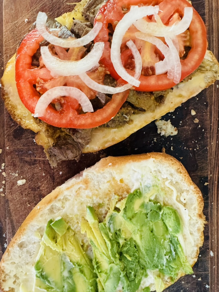 Sliced tomatoes and onions on top of steak, beans, and cheese on a torta. Smashed avocado on top of mayo on other half of bread