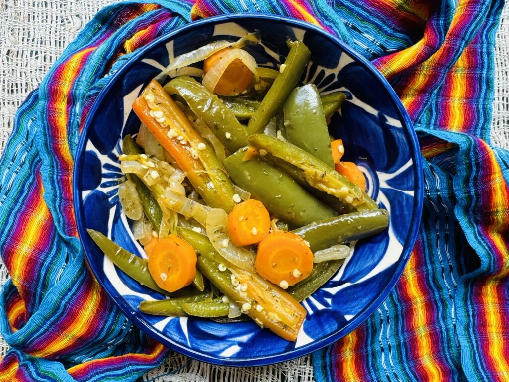 Pickled jalapeños, onions, and carrots in a blue pottery bowl with a colorful Mexican rebozo in the background