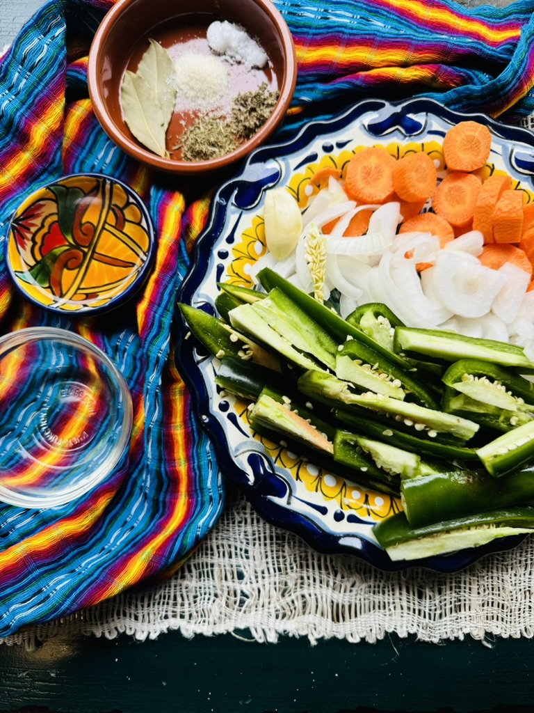 Sliced jalapeños, carrots and onions on a Mexican pottery plate, vinegar, oil and spices are in 3 small bowls.