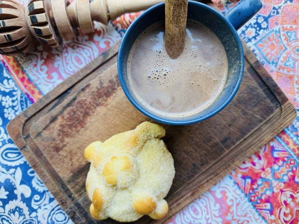 Mexican hot chocolate in a blue mug with pan de muerto and a molinillo