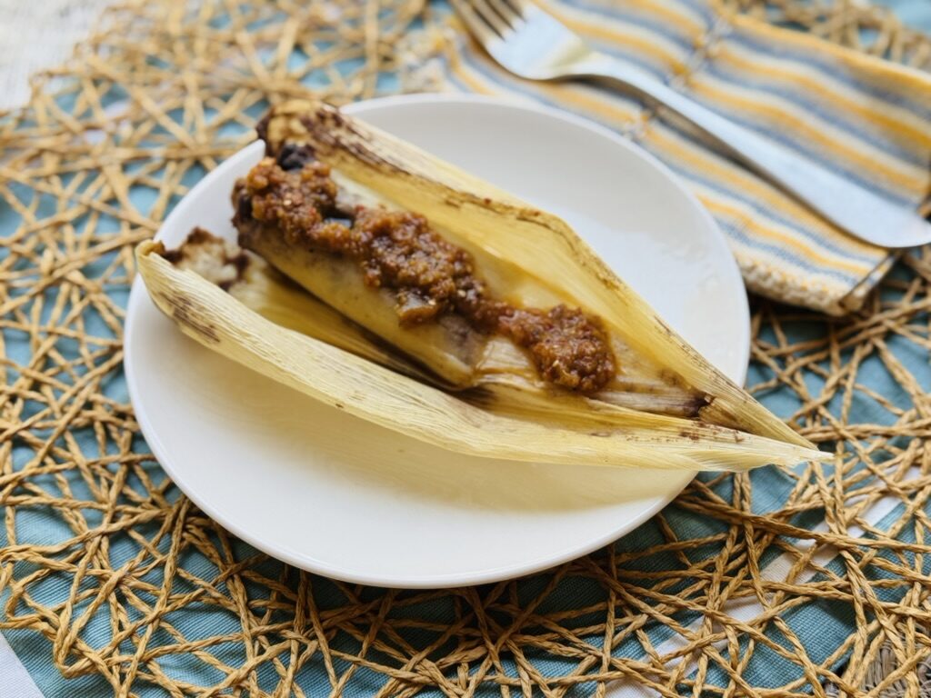 Corn husk tamal on a plate, topped with roasted red salsa